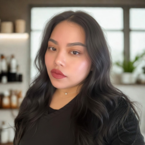 Portrait of a woman with dark wavy hair wearing a black top, looking at the camera in a bright room with blurred shelves in the background.